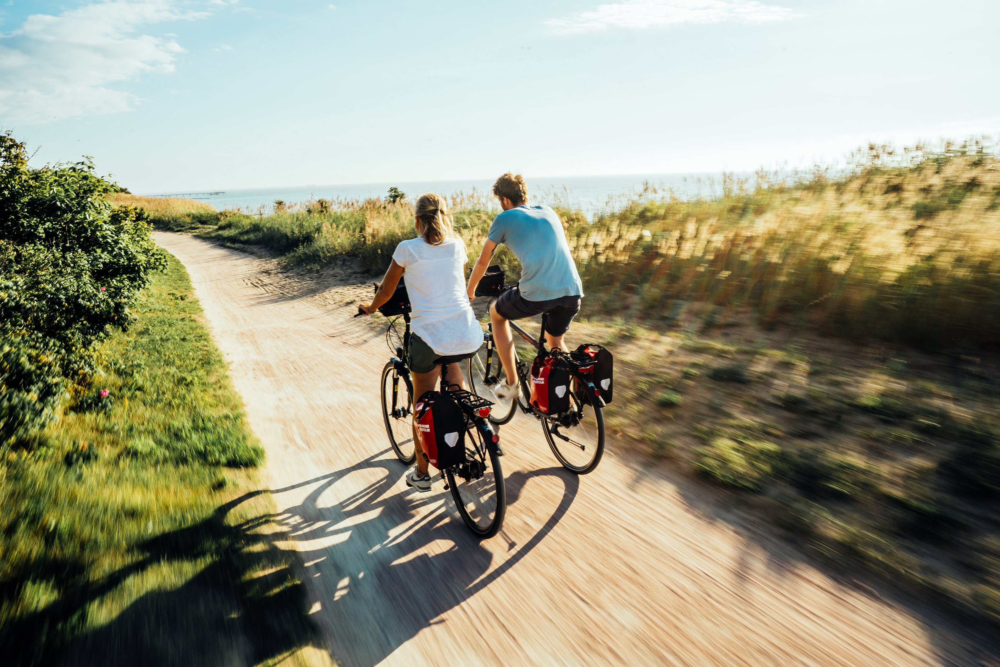 Zwei Fahrradfahrer fahren auf einem Sandweg entlang der Küste. Man sieht die Fahrradfahrer nur von hinten